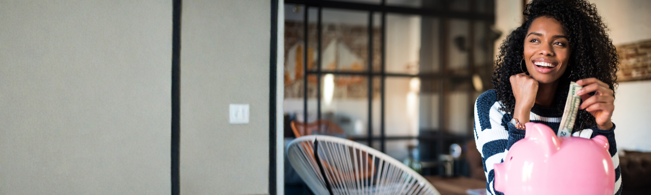 A smiling woman inserting a dollar bill into a pink piggy bank in a modern living room, representing spring savings goals.